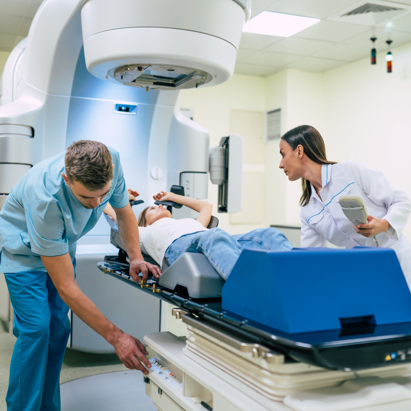 Healthcare professionals preparing a patient for a medical imaging scan, with one technician operating equipment controls while another assists the patient positioned on the scanning table beneath a large white medical imaging machine.