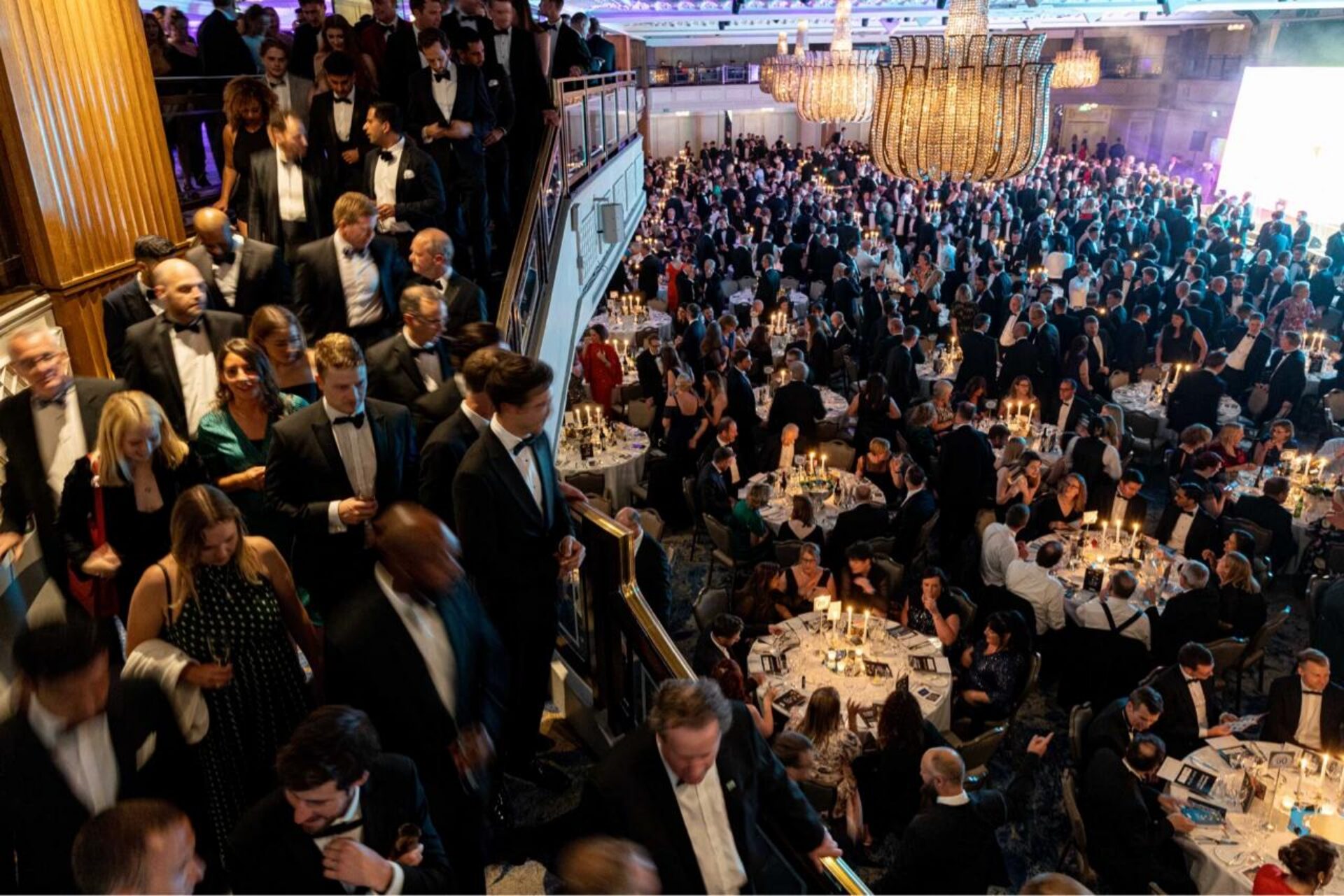 A formal black-tie gala event in an elegant ballroom with hundreds of attendees in evening wear gathered around dining tables on the main floor, while others socialize on a mezzanine level above, all under ornate crystal chandeliers.