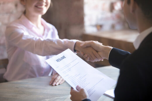 A job candidate in a pink shirt shakes hands with an interviewer in a dark suit who is holding her resume, suggesting a successful job interview conclusion.