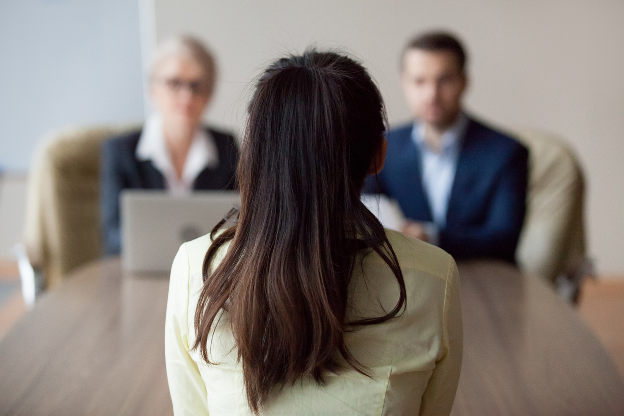 A woman with long dark hair sits facing two interviewers across a table in what appears to be a job interview setting, with the interviewers slightly blurred in the background.
