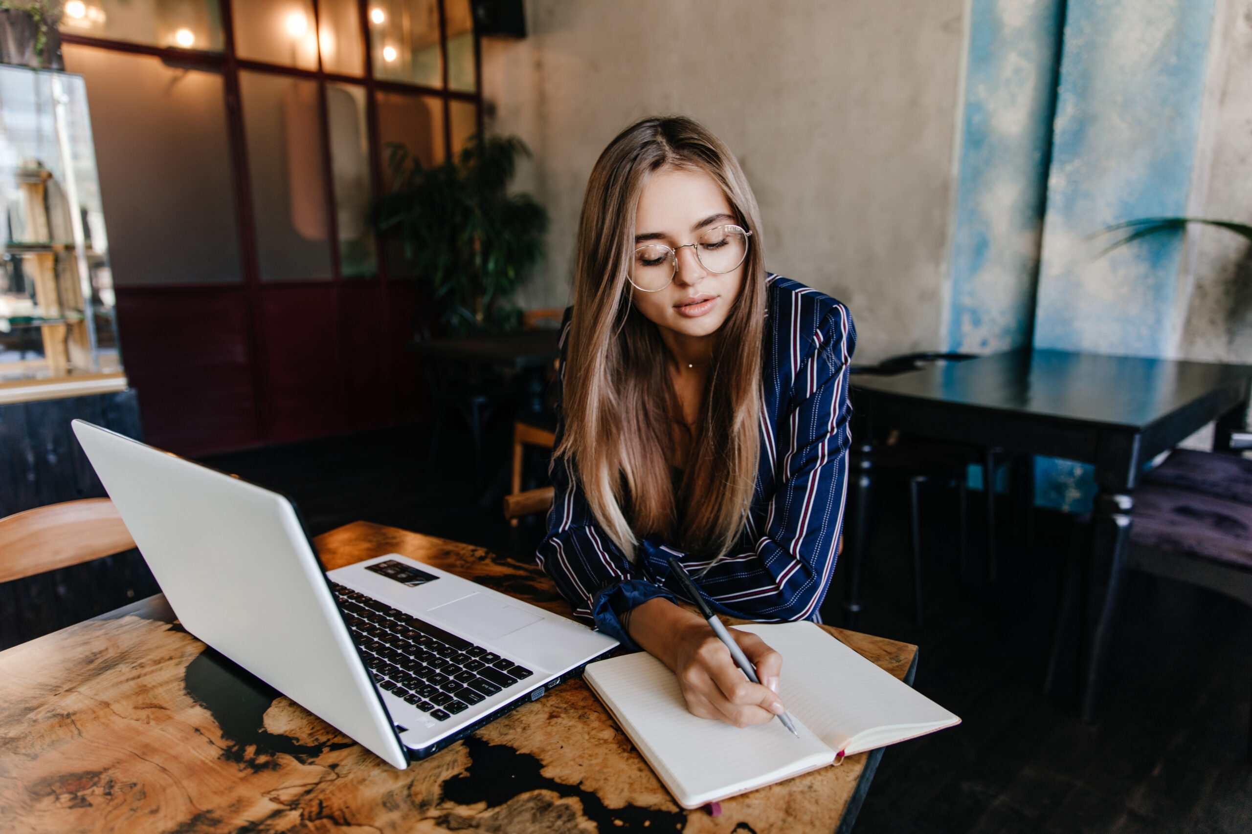 A woman with glasses and long brown hair wearing a blue striped shirt sits at a wooden table, writing in a notebook while working on a laptop in a modern cafe or office space with warm lighting.