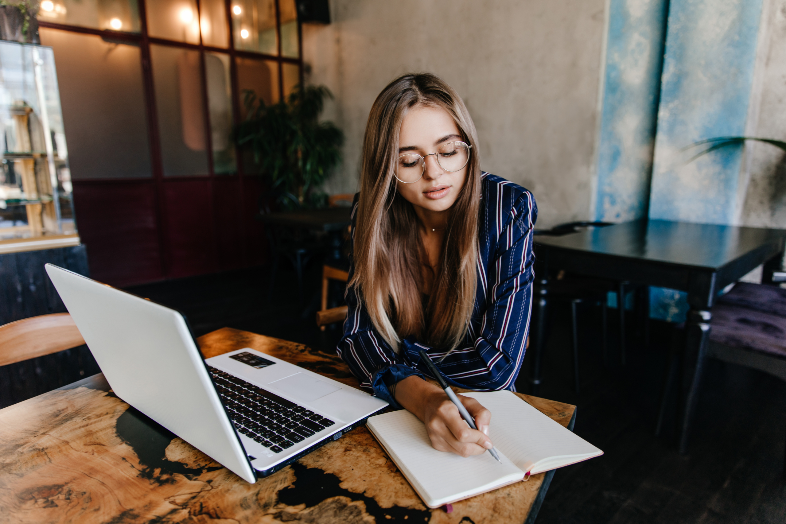 A woman with glasses and long brown hair wearing a blue striped shirt sits at a wooden table, writing in a notebook while working on a laptop in a modern cafe or office space with warm lighting.