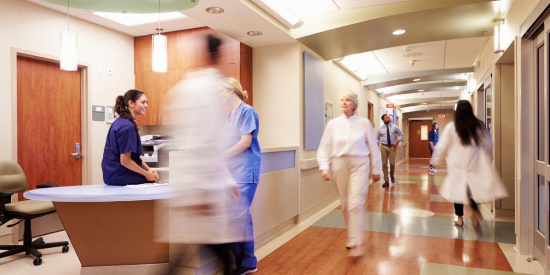 A busy hospital corridor with medical staff in scrubs moving quickly past a nurses' station, creating motion blur that emphasizes the fast-paced healthcare environment.