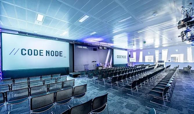 A modern conference auditorium with tiered seating facing a large projection screen displaying the "CODE NODE" logo, featuring contemporary lighting and professional presentation setup.