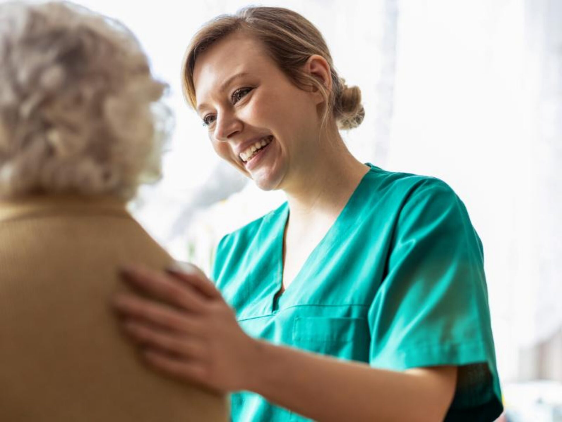 A smiling healthcare worker in green scrubs provides compassionate care to an elderly patient with curly gray hair in a bright medical facility.