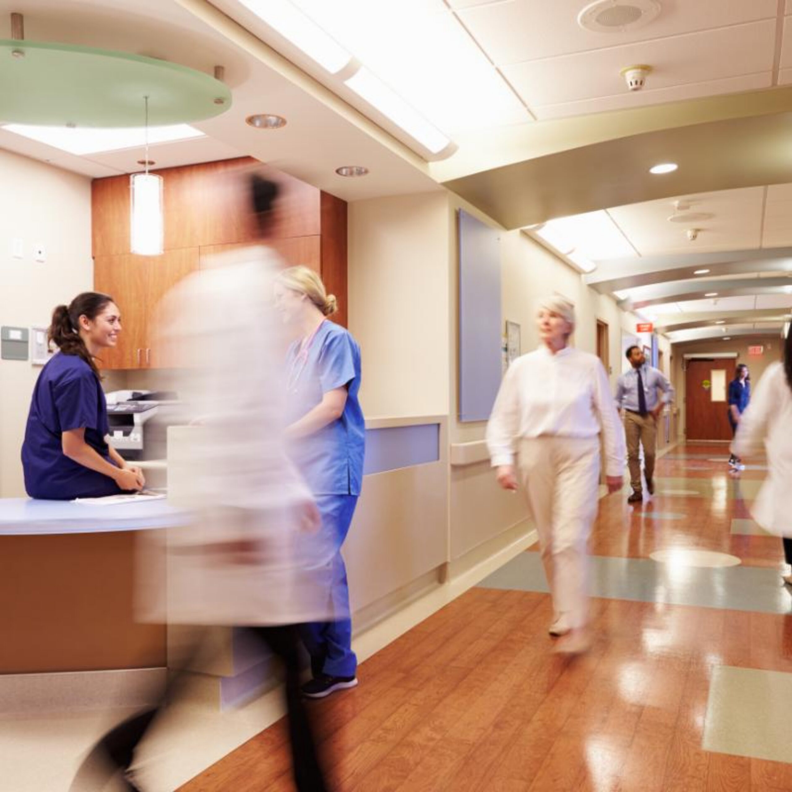 Healthcare workers in scrubs move through a busy hospital corridor, with some figures appearing blurred due to their rapid movement while one nurse remains stationary at a reception desk.